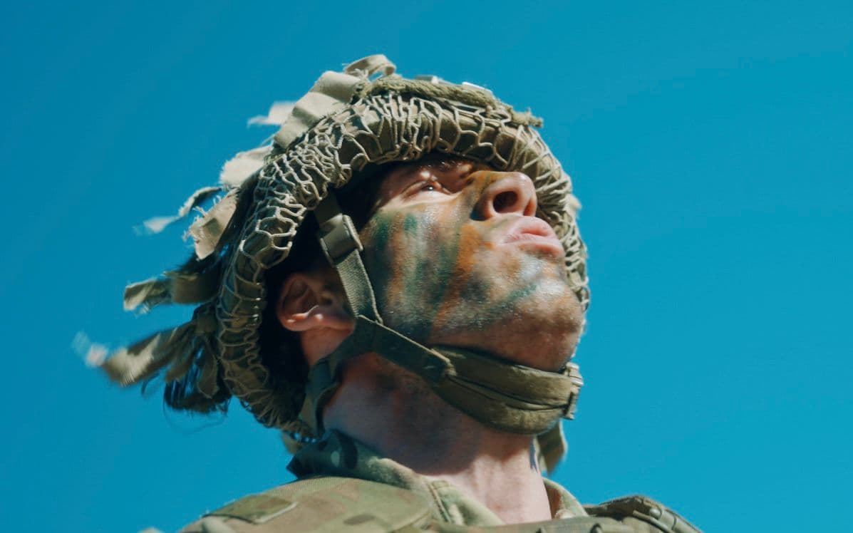 Tristan Alexander as a soldier with camouflage face paint looking upward against blue sky