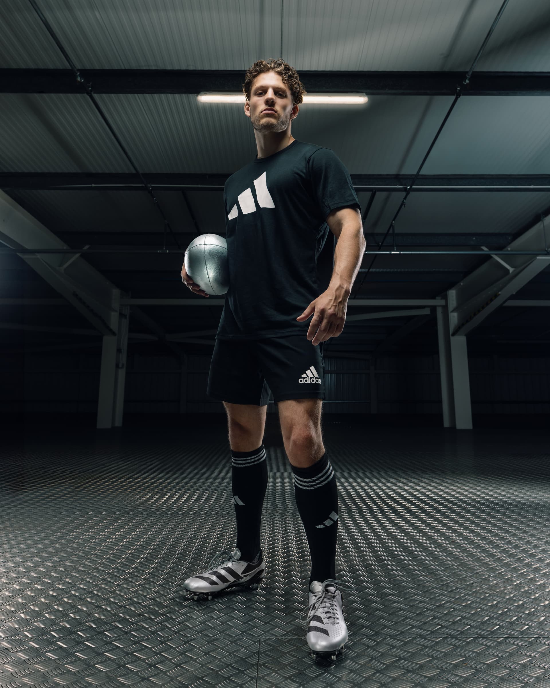 Tristan Alexander in full Adidas kit holding a football in a dark industrial warehouse